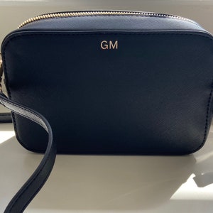 May include: Black leather crossbody bag with a gold zipper and a gold metal clasp. The bag has a gold monogram "GM" on the front.