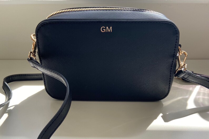 May include: Black leather crossbody bag with a gold zipper and a gold metal clasp. The bag has a gold monogram "GM" on the front.