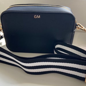 May include: A black crossbody bag with a gold zipper and a white and black striped strap. The bag has the initials "GM" embossed on the front.