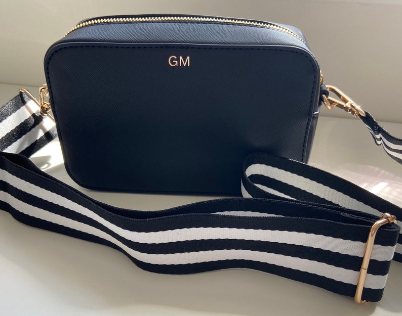 May include: A black crossbody bag with a gold zipper and a white and black striped strap. The bag has the initials "GM" embossed on the front.