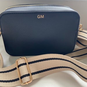 May include: A black camera bag with a gold zipper and a gold strap buckle. The bag has a cream and black striped adjustable strap and the initials "GM" are embossed in gold on the front of the bag.