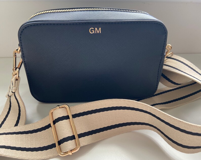 May include: A black camera bag with a gold zipper and a gold strap buckle. The bag has a cream and black striped adjustable strap and the initials "GM" are embossed in gold on the front of the bag.