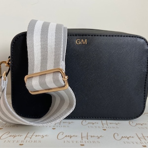 May include: Black camera bag with a white and gray striped adjustable strap. The bag has a gold metal buckle and the initials "GM" are embossed on the front.