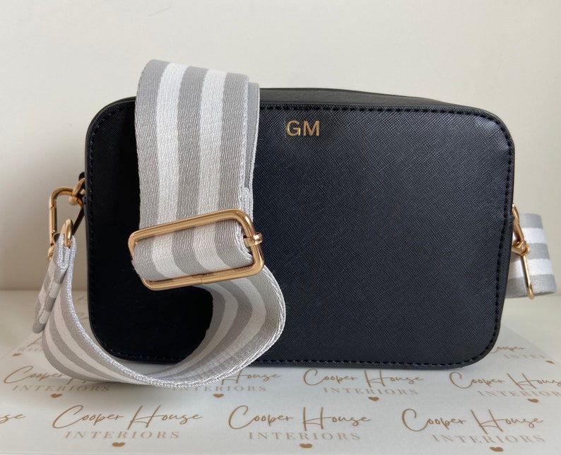 May include: Black camera bag with a white and gray striped adjustable strap. The bag has a gold metal buckle and the initials "GM" are embossed on the front.