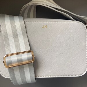 May include: A light gray crossbody bag with a white and gray striped strap. The bag has a gold buckle and the initials "JB" are embossed on the front.