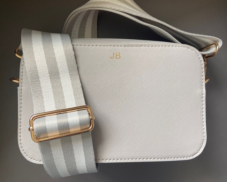 May include: A light gray crossbody bag with a white and gray striped strap. The bag has a gold buckle and the initials "JB" are embossed on the front.