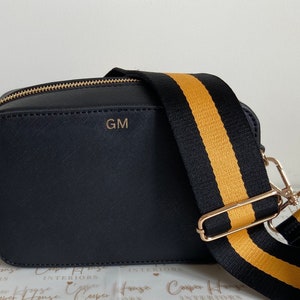 May include: A black crossbody bag with a gold zipper and a black and gold striped strap. The bag has the initials "GM" embossed on the front.