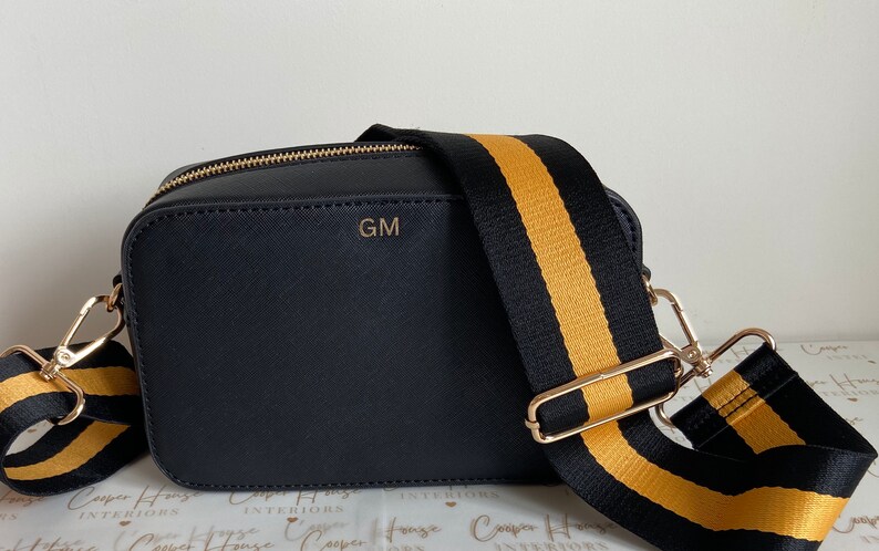 May include: A black crossbody bag with a gold zipper and a black and gold striped strap. The bag has the initials "GM" embossed on the front.