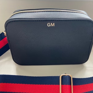 May include: A black crossbody bag with a gold zipper and strap. The bag has a red and blue striped strap and the initials "GM" are embossed on the front.