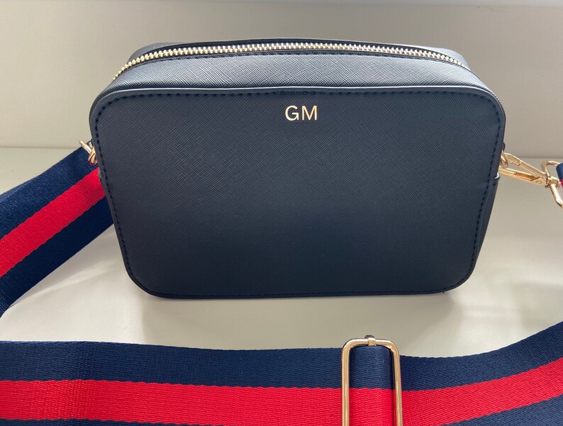 May include: A black crossbody bag with a gold zipper and strap. The bag has a red and blue striped strap and the initials "GM" are embossed on the front.