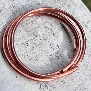 May include: A coiled copper wire bracelet featuring a smooth, metallic finish. The design consists of multiple overlapping circular loops of polished copper wire, creating a continuous, layered appearance with rounded ends visible on the outer coil.