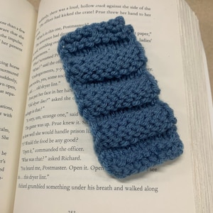 May include: A close-up of a blue knitted bookmark with a textured pattern. The bookmark is lying on top of an open book.