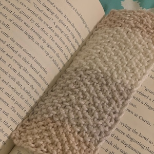 May include: A close-up of a knitted beige and white bookmark with a chevron pattern. The bookmark is lying on top of an open book with text about a story.