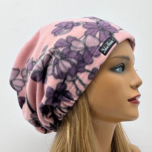 May include: A pink fleece beanie with a floral pattern in shades of purple and black. The beanie has a soft, slouchy fit and features a small black label with white text that reads "Deluxe Beanie".