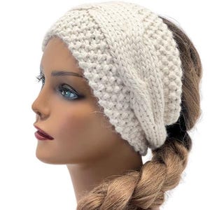 May include: Cream-colored knitted headband with a cable knit design. The headband is made of a thick, textured yarn and is worn on a mannequin with a blonde braid. The headband covers the forehead and ears.
