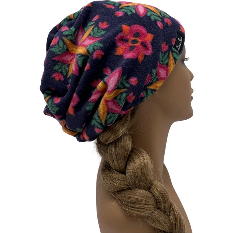 May include: A navy blue fleece beanie hat with a floral pattern in pink, orange, yellow, and green. The hat has a relaxed fit and a small black label with white text. The hat is shown on a mannequin head with a blonde braid.