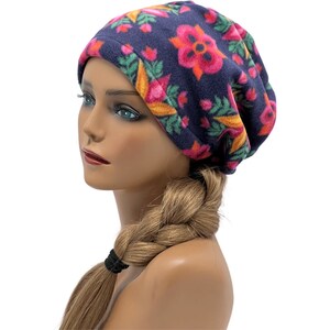 May include: A navy blue fleece beanie hat with a floral pattern in shades of pink, orange, and green. The hat is displayed on a mannequin head with a blonde braid. The beanie is a casual accessory.