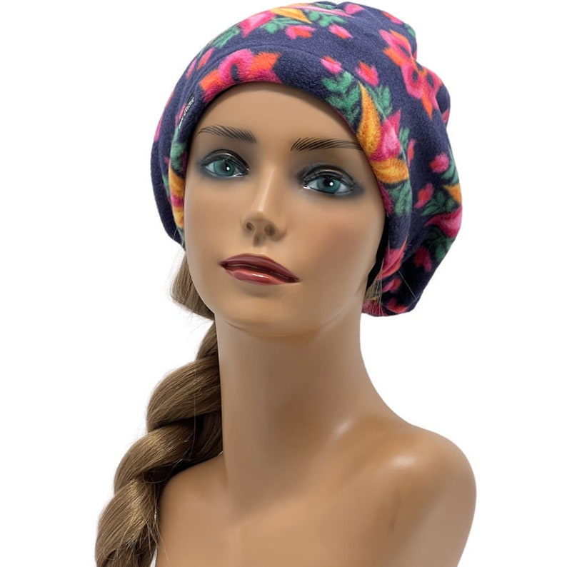 May include: A navy blue fleece hat with a floral pattern in pink, orange, and green. The hat is a beret style and is displayed on a mannequin head. The hat is designed to keep the head warm.