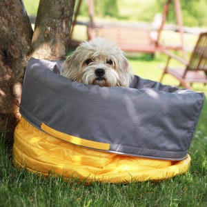 May include: A small dog rests in a grey and yellow pet bed in a garden setting. The bed features a grey, padded upper section and a yellow, quilted base. The dog has light-coloured, fluffy fur. The bed is placed on grass.