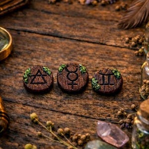 Peut inclure: Une table en bois rustique présente trois médaillons ronds et bruns gravés de symboles astrologiques. La scène comprend une bougie allumée, des parchemins, une loupe et de petits bocaux en verre, évoquant un thème mystique.