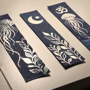 Peut inclure: Trois marque-pages bleu marine avec des motifs argentés. L'un représente une méduse, un autre un croissant de lune et le troisième un symbole Om. Chaque marque-page a un motif botanique unique en argent.