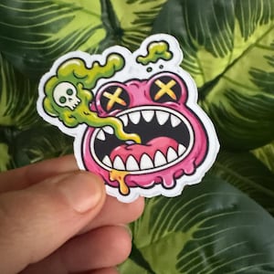 Peut inclure: Autocollant de grenouille rose de dessin animé avec une grande bouche ouverte, des yeux croisés jaunes et un nuage de fumée verte avec un crâne. L'autocollant a une bordure blanche et est tenu par une main.