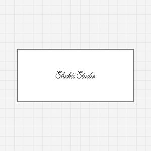 May include: A white rectangular card with the words "Shakti Studio" written in elegant cursive script. The text is centered on the card, which is set against a light gray grid background. The overall design is simple and clean.