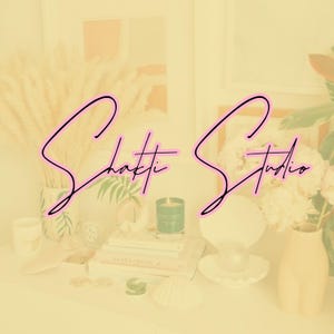 May include: A decorative shelf with the words "Shakti Studio" in a pink script. The shelf displays a vase of dried wheat, books, a candle, a seashell, and a decorative lamp.