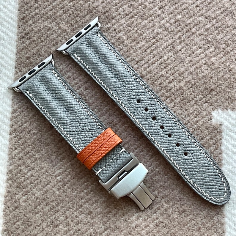 May include: Grey leather watch strap with white stitching and a stainless steel clasp. The strap has an orange leather loop and is designed for an Apple Watch. The watch strap is shown on a textured, neutral-coloured surface.