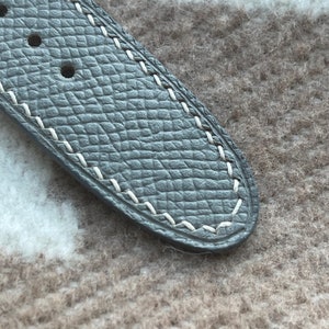 May include: Close-up of a grey leather watch strap with white stitching. The strap has a textured surface and three holes for adjustment. The stitching is visible along the edges of the strap.
