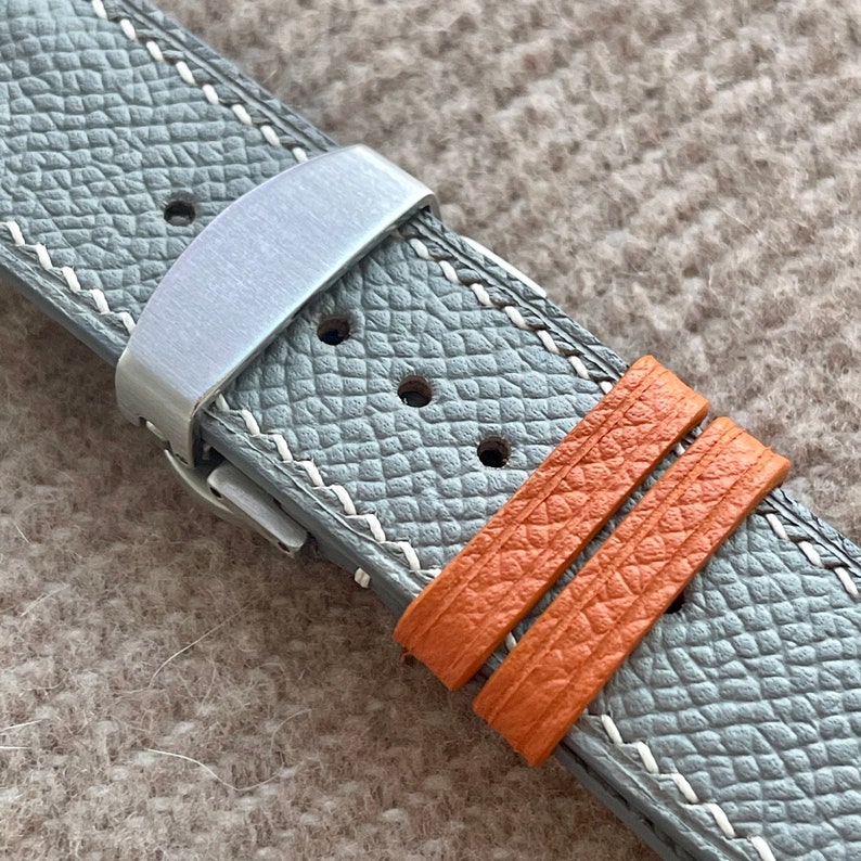 May include: Close-up of a grey leather watch strap with white stitching and a silver clasp. Two orange leather keepers are visible. The strap has a textured surface.