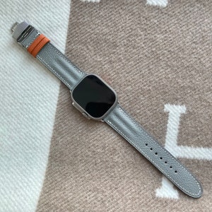 May include: A silver smartwatch with a grey leather strap and orange detail. The watch has a rectangular black screen. The strap is stitched with white thread and has adjustment holes. The watch is displayed on a beige and white patterned surface.
