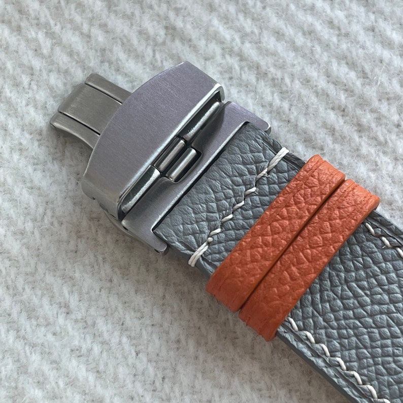 May include: Close-up of a grey leather watch strap with orange accents and white stitching. The strap features a stainless steel clasp. The leather has a textured appearance.