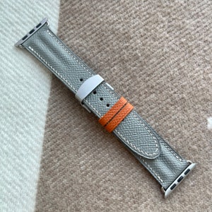 May include: A grey leather watch strap with white stitching and silver fittings. The strap has a textured surface and an orange accent strap. The watch strap is designed for a smartwatch.
