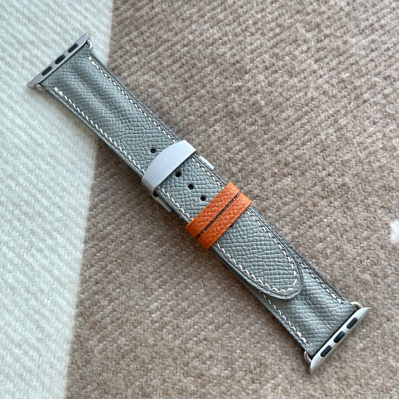 May include: A grey leather watch strap with white stitching and silver fittings. The strap has a textured surface and an orange accent strap. The watch strap is designed for a smartwatch.