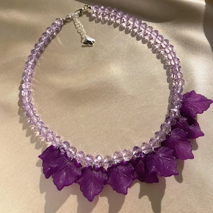 Puede incluir: Un collar que presenta una hilera de cuentas de cristal facetado en color violeta claro y una serie de dijes con forma de hoja en color violeta oscuro mate en la parte inferior. Incluye un cierre de mosquetón plateado y una cadena extensora.