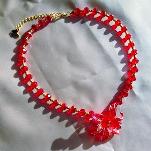 Puede incluir: Un collar de cuentas rojas con una cadena dorada y un colgante de flor roja. El collar presenta pequeñas cuentas rojas redondas ensartadas con metal dorado. El colgante de flor es de un rojo vibrante.