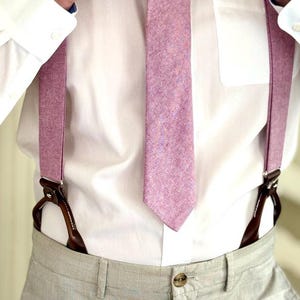 May include: A person adjusting a light purple tie, with matching suspenders. The tie and suspenders are made of a textured fabric. The person is wearing a white dress shirt and light gray pants.