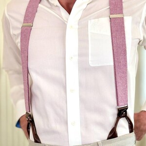 May include: A close-up of a person wearing a white button-down shirt and light gray pants. The person is wearing pink suspenders with silver metal clasps and brown leather attachments.