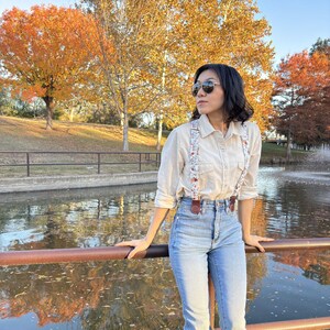 Women’s floral suspenders with leather magnetic clasp attachments, styled with blue jeans and a cream button-up shirt beside a pond with fall foliage.