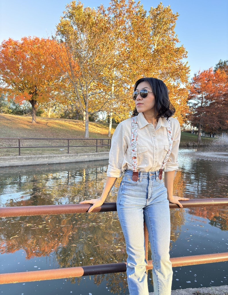 Women’s floral suspenders with leather magnetic clasp attachments, styled with blue jeans and a cream button-up shirt beside a pond with fall foliage.
