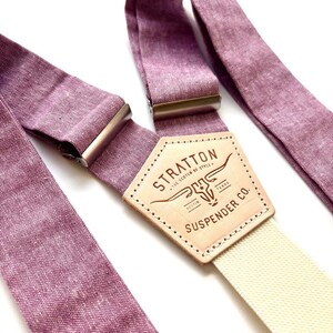 May include: Close-up of purple suspenders with a cream-colored strap. The suspenders have metal clasps and a leather patch with the text "STRATTON SUSPENDER CO. AUSTIN TEXAS". The suspenders are made of a textured fabric.