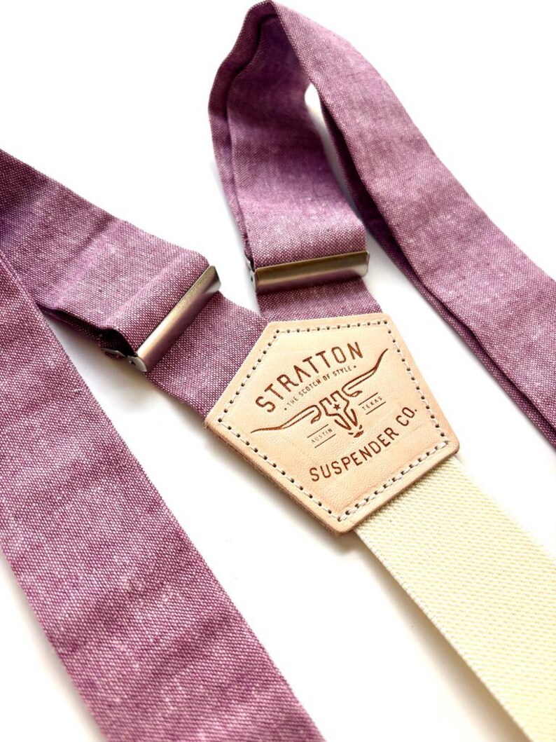 May include: Close-up of purple suspenders with a cream-colored strap. The suspenders have metal clasps and a leather patch with the text "STRATTON SUSPENDER CO. AUSTIN TEXAS". The suspenders are made of a textured fabric.