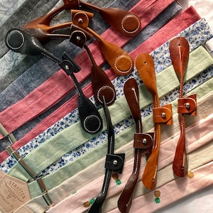 May include: Assortment of leather straps with metal hardware and pineapple designs. The straps are in various colors, including brown, black, and white. The straps are arranged on a background of patterned fabric.