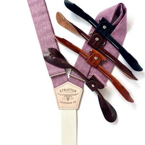 May include: A close-up of a pair of lavender suspenders with leather attachments in various shades of brown and black. The suspenders have a cream-colored elastic band and a leather patch with the text "STRATTON SUSPENDER CO."