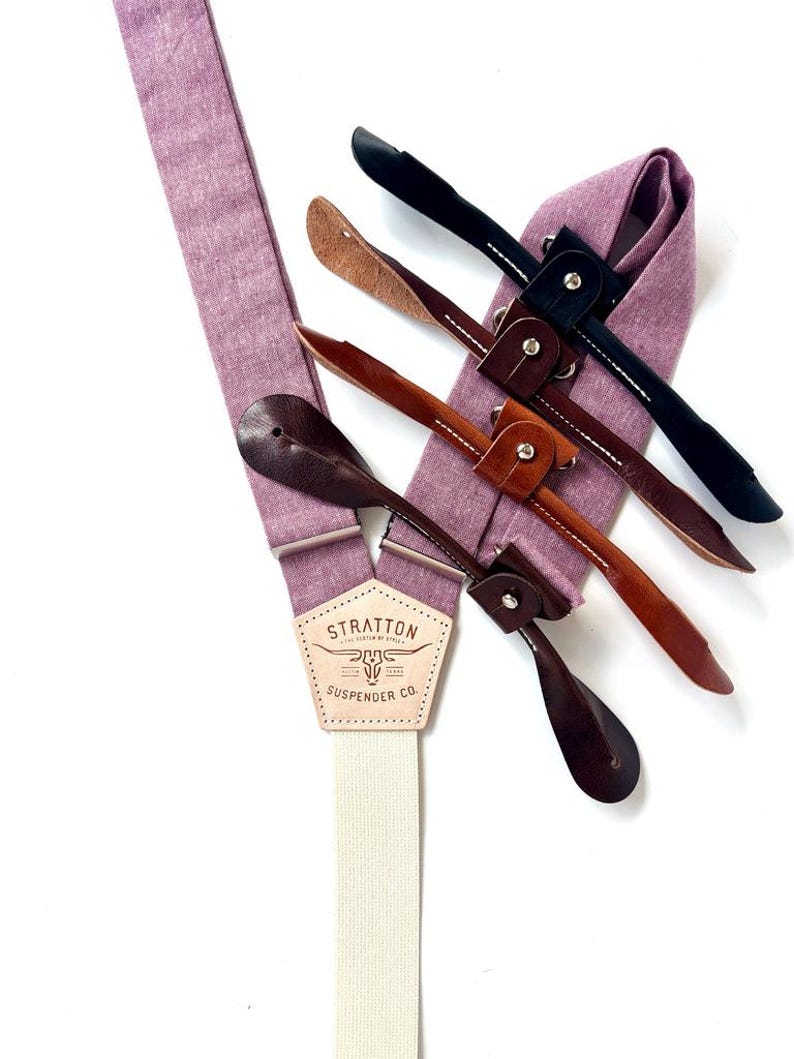 May include: A close-up of a pair of lavender suspenders with leather attachments in various shades of brown and black. The suspenders have a cream-colored elastic band and a leather patch with the text "STRATTON SUSPENDER CO."