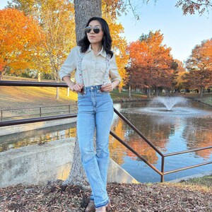Full-body view of women’s floral magnetic clasp suspenders worn with light blue jeans, a cream shirt, and brown boots in an autumn park setting.