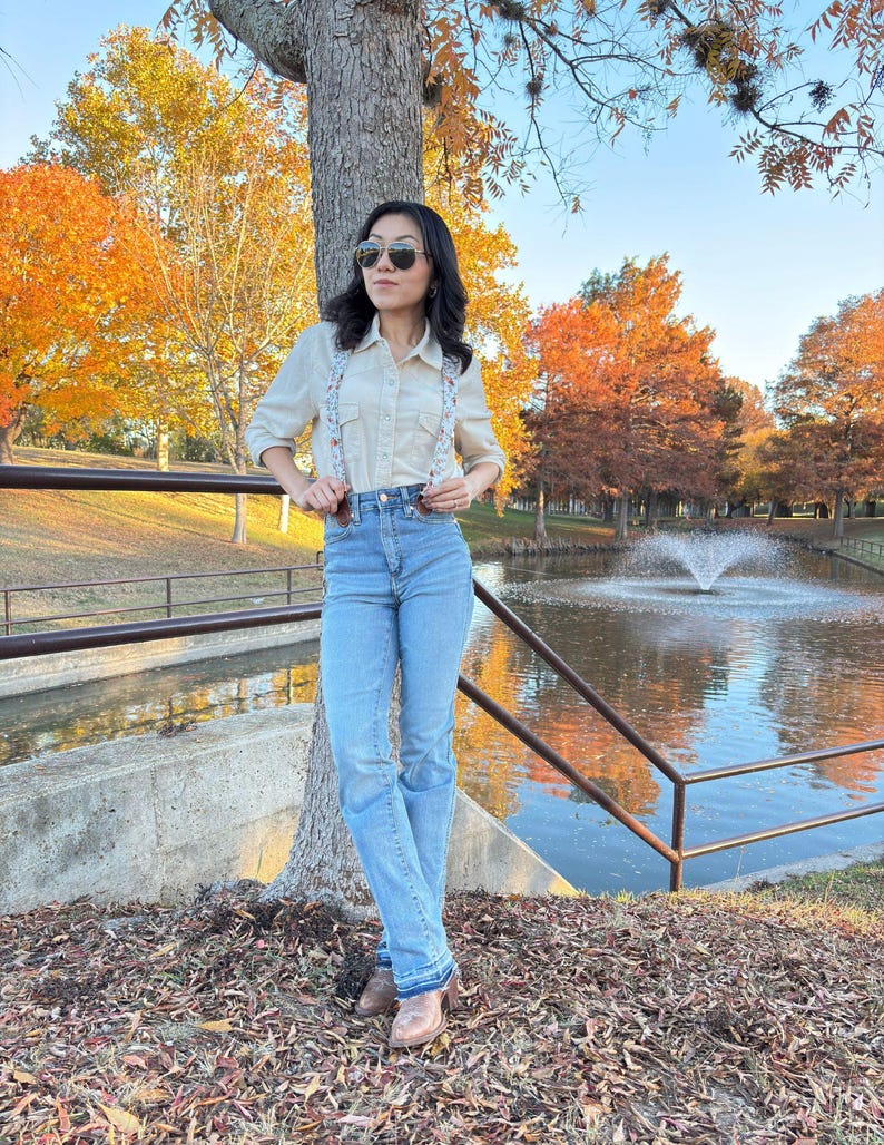 Full-body view of women’s floral magnetic clasp suspenders worn with light blue jeans, a cream shirt, and brown boots in an autumn park setting.