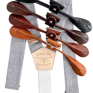 May include: Gray linen suspenders with a leather buckle and adjustable straps. The buckle features a brown leather patch with the text "Stratton Suspender Co." and a bull's head logo.
