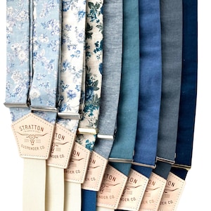 Puede incluir: Un conjunto de 10 tirantes en varios tonos de azul, incluyendo patrones florales y colores sólidos. Los tirantes están hechos de algodón y tienen una correa de cuero con el texto "Stratton Suspender Co."